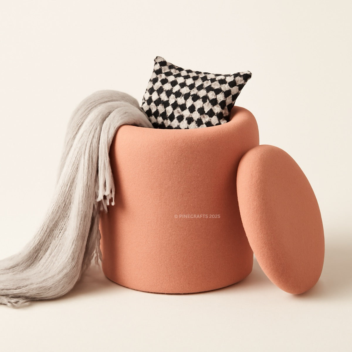 Cylindrical pink ottoman with a checkered pillow and gray scarf on a light background