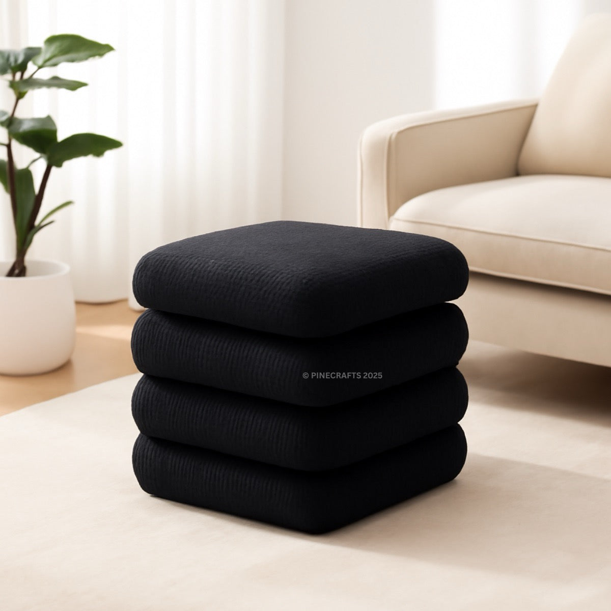Stack of black cushions on a light-colored floor with a beige sofa and plant in the background.