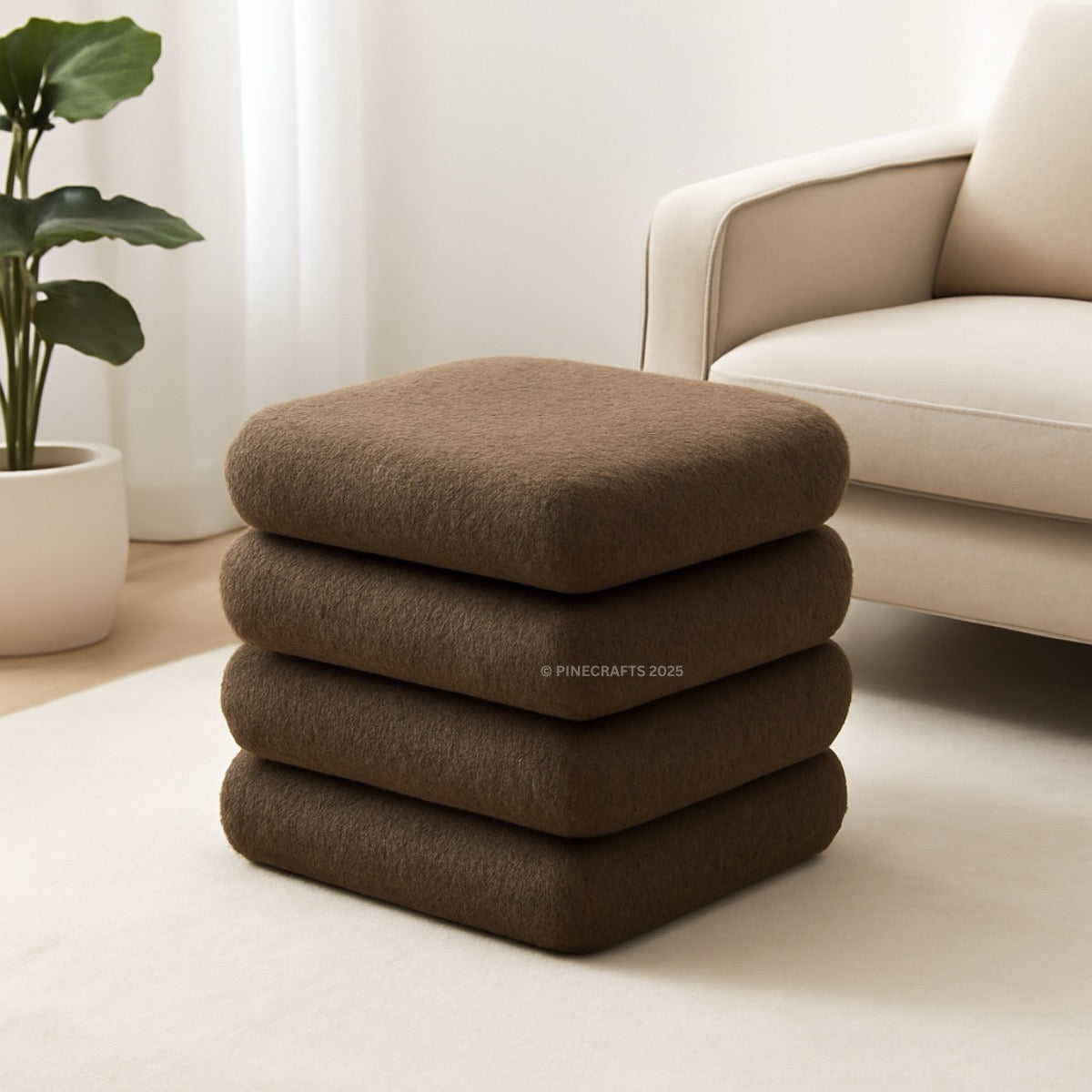 Stack of brown cushions on a light-colored floor with a beige armchair and plant in the background.