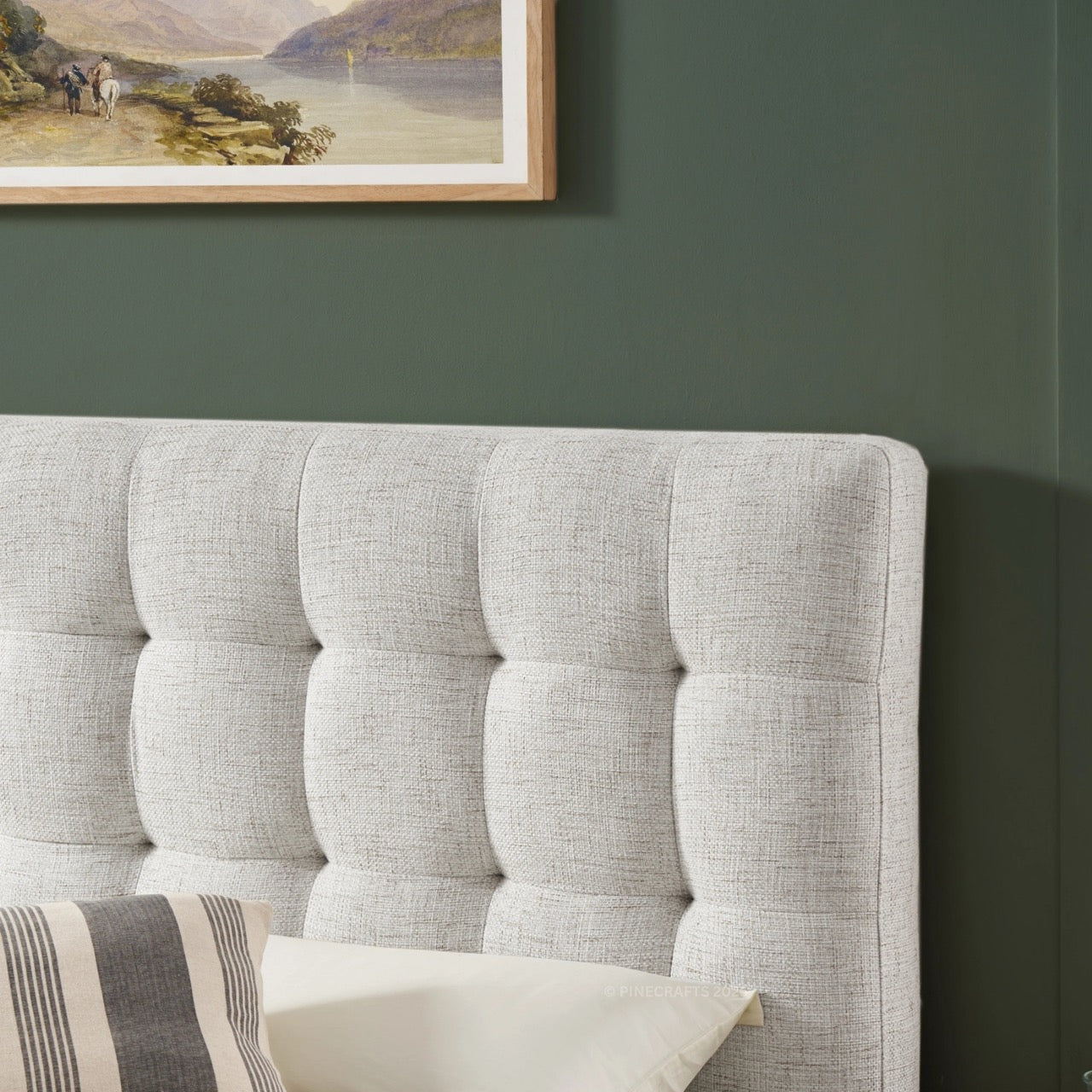 Gray tufted headboard with pillows against a green wall with a framed landscape painting.