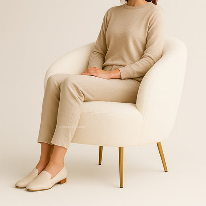 Person wearing a beige sweater and pants sitting on a white chair with gold legs.