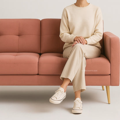 Person sitting on a pink couch wearing a beige sweater and pants with white sneakers.