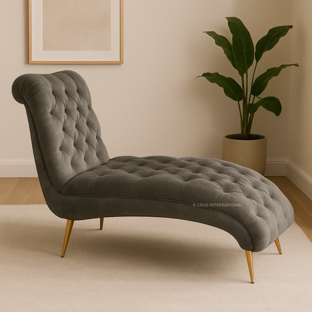 Gray tufted chaise lounge with gold legs in a room with a plant and framed picture.