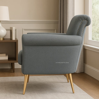 Gray armchair with gold legs in a room with a lamp and books on a side table.