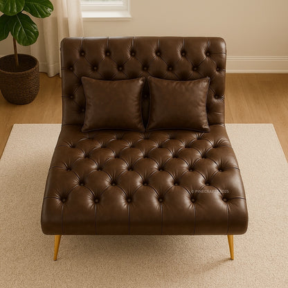 Brown leather tufted chaise lounge with two pillows in a room with a plant and window.